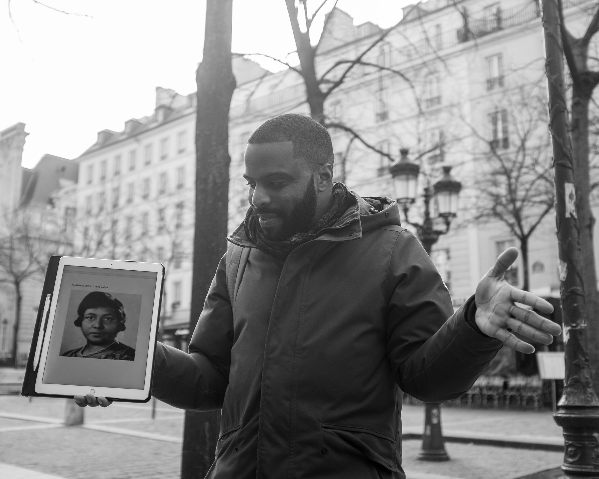 Black and white photograph of Kévi Donat. He's standing across the street from an old building, presumably in Paris, and he's facing the camera with arms outstretched. In his right hand is a tablet with the picture of a Black woman. He's looking down at the tablet.