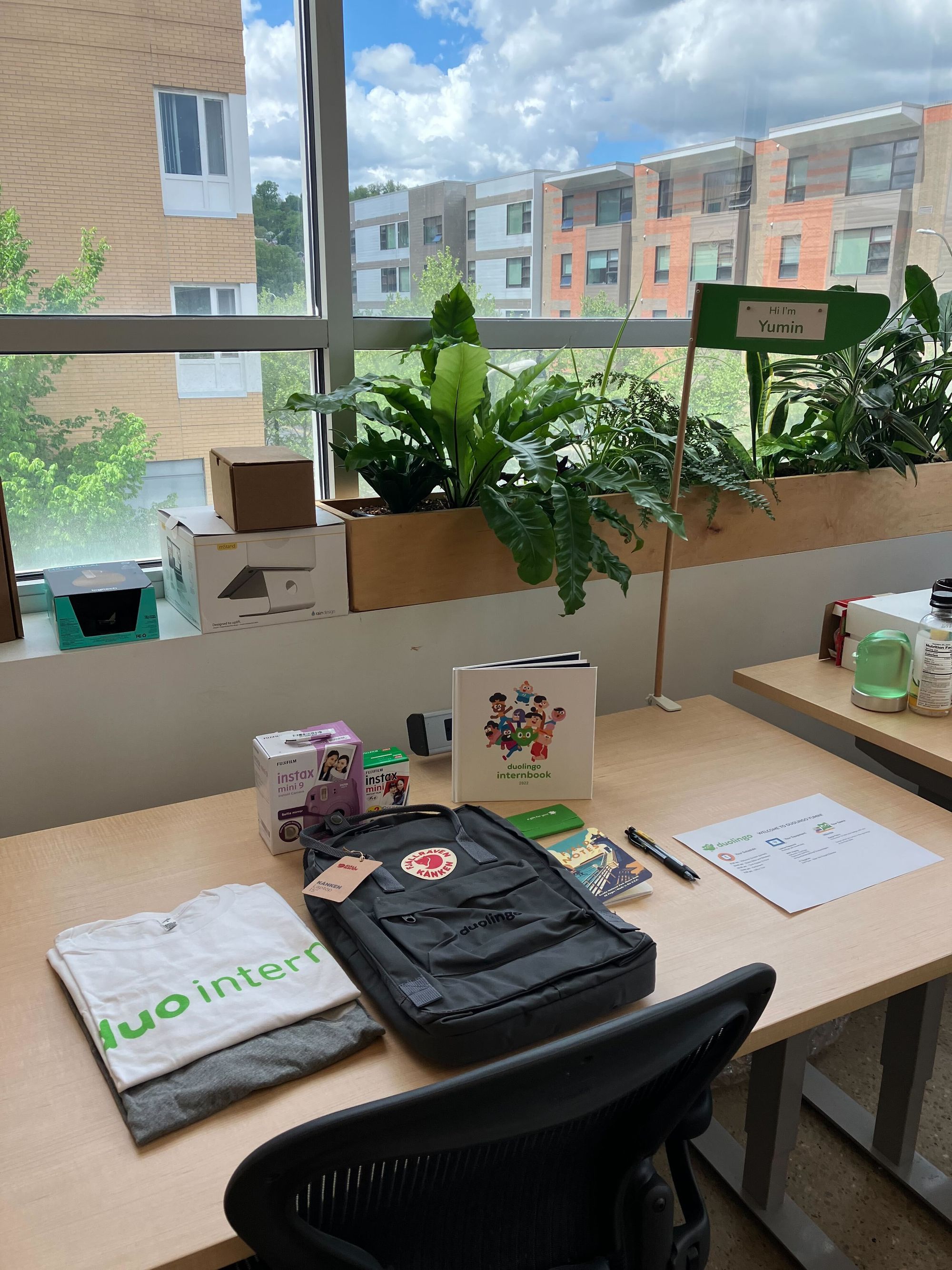 A sunny desk surrounded by plants, featuring a backpack, a t-shirt that says "duointern", a book, and a polaroid camera