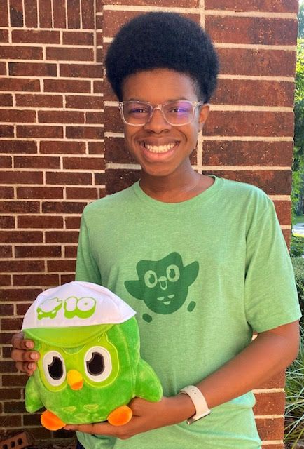 Image of Colton Holiday, smiling, wearing a Duolingo t-shirt. He's holding a Duolingo plushie