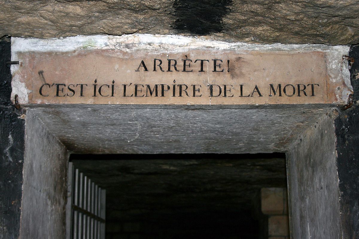 The Catacombs entrance is a dark-colored wall with a warning sign above a door opening that reads “Arrête! C’est ici l’empire de la mort.” This translates to “Stop! Here begins the empire of death.” Photo Credit: Gaspard Duval