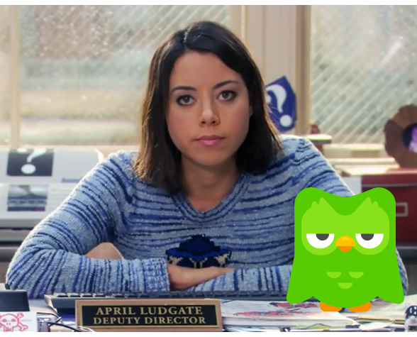 Still shot from "Parks and Recreation" of April Ludgate sitting at her desk looking unamused. The Duolingo owl, looking similarly unimpressed, is photoshopped onto her desk next to her.