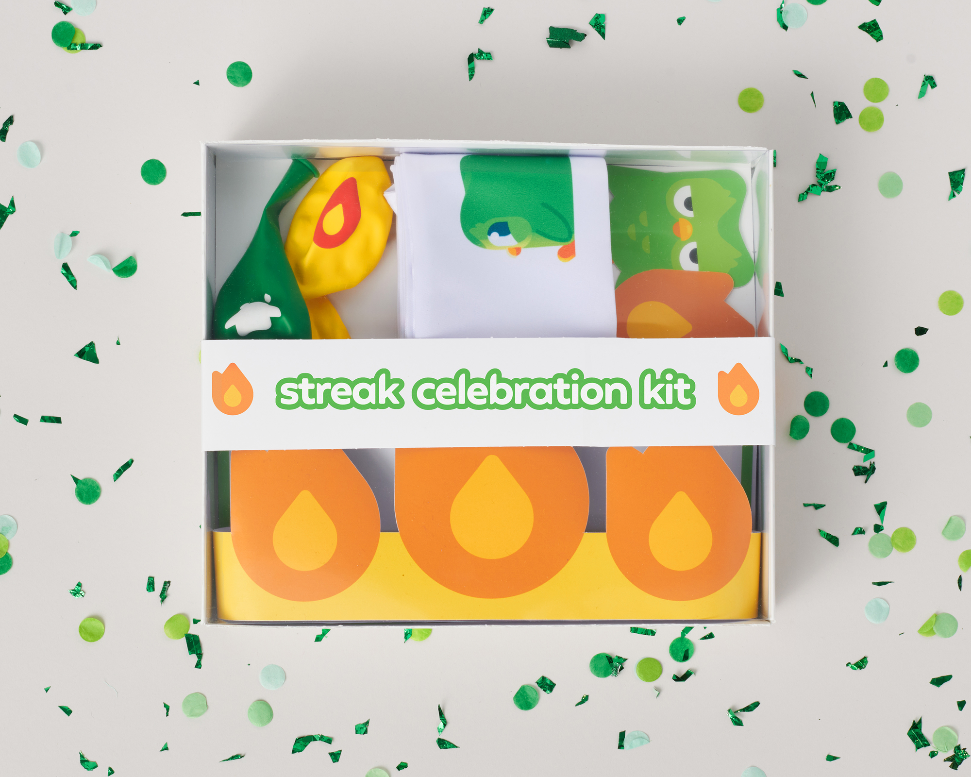 A picture of the Duolingo Streak Celebration Kit. A box with party items, like balloons, a sash, and a Duo banner, are in a box surrounded by confetti
