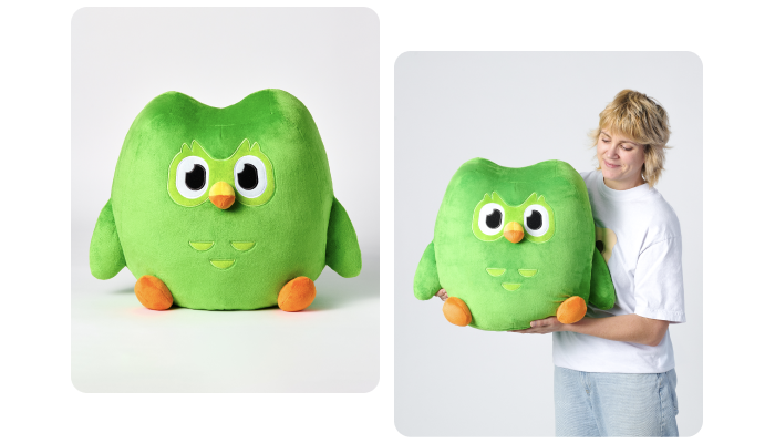 A very large version of the Duolingo plushie. He is large and round, and you can see from the blonde adult woman hugging him in her arms that he is almost the size of her torso.