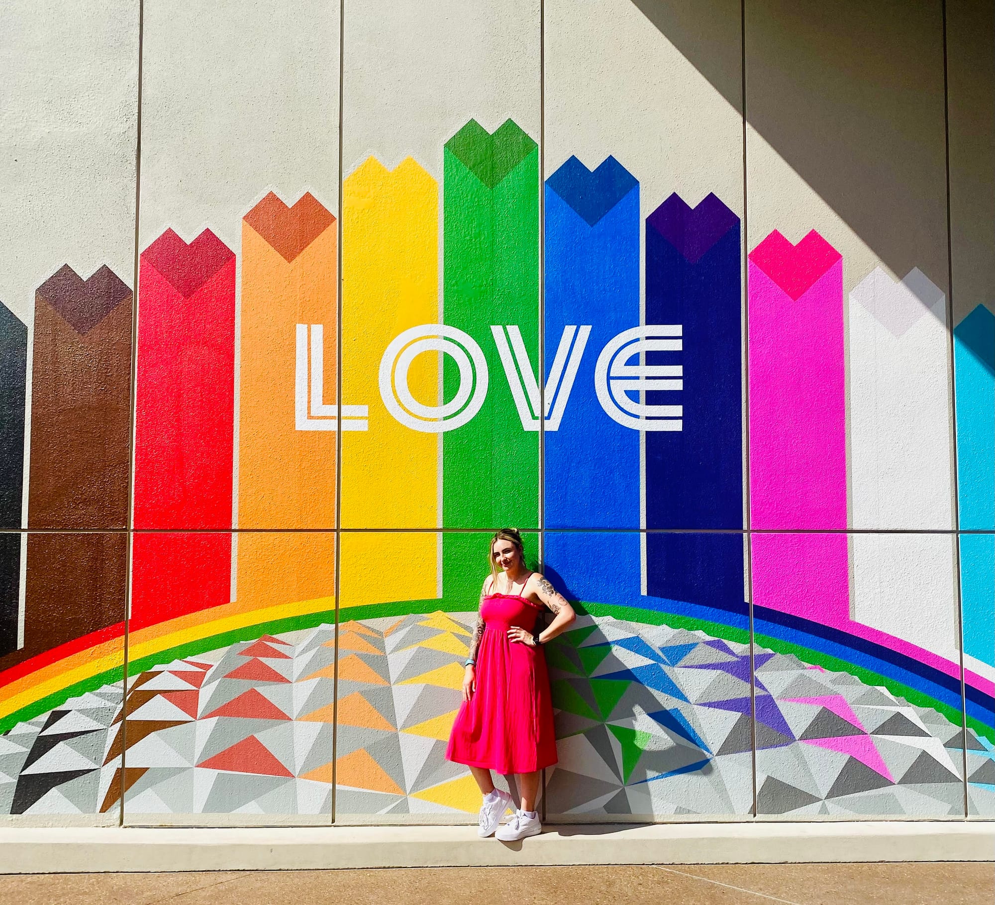 Photo d’Adèle debout devant un immeuble avec une fresque arc-en-ciel sur laquelle on peut lire le mot « LOVE »
