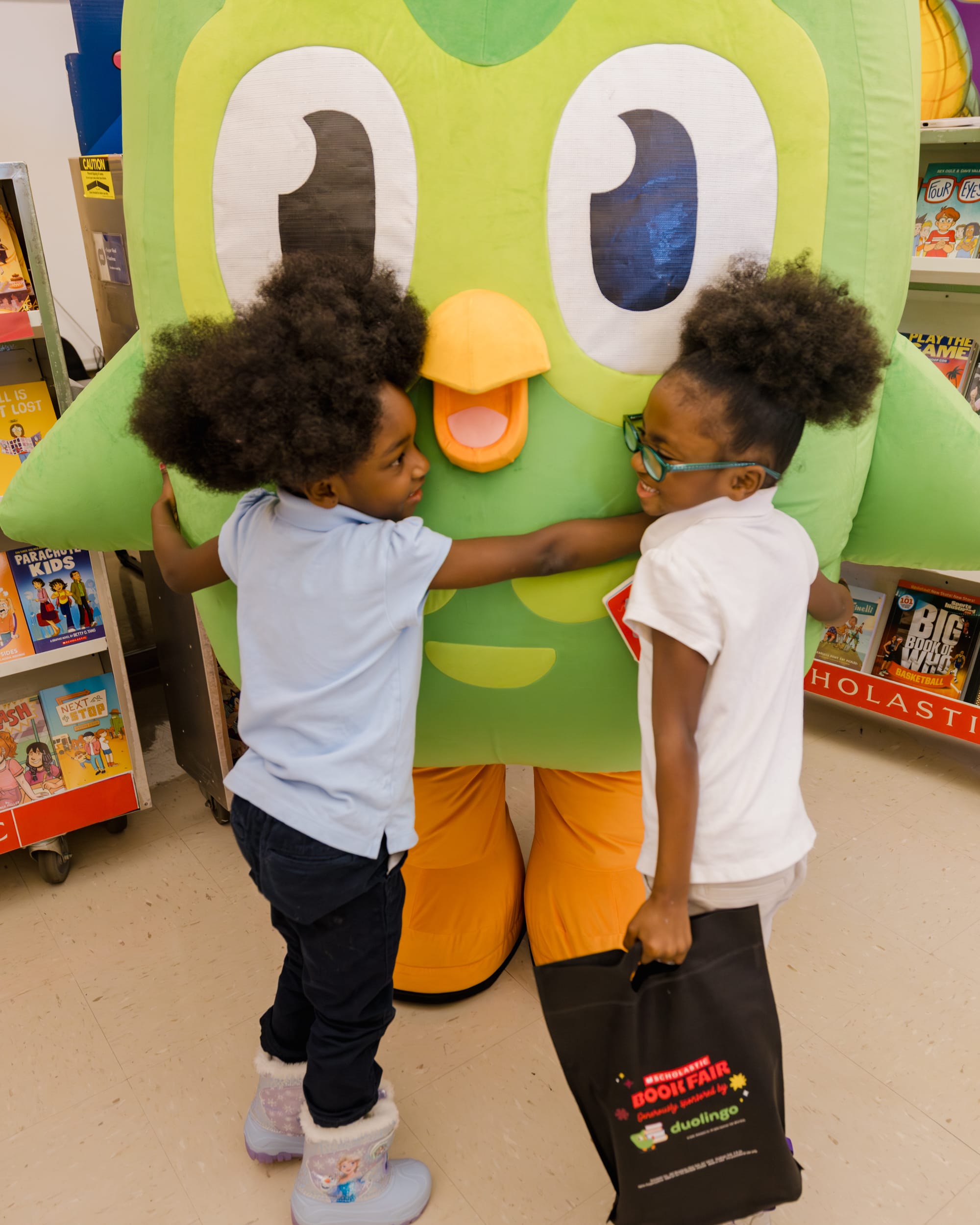 Two children hugging Duo the Owl. One of the children holds a Duolingo and Scholastic Book Fair tote bag.
