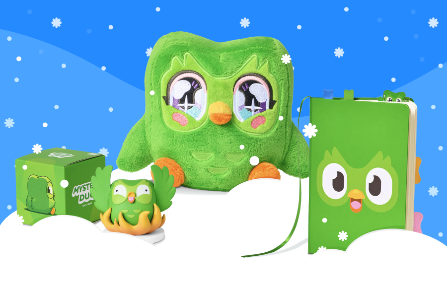 Ein Anime-Kuschel-Duo, ein Duolingo-Notizbuch und eine Überraschungsbox mit einer Duo-Figur daneben auf einem Schneehaufen